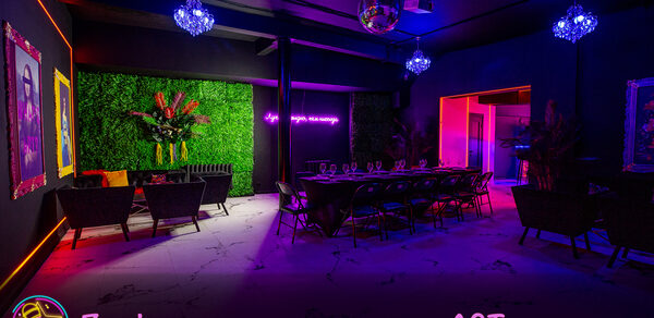Белый неоновый лофт-зал. Зал для мероприятий с мебелью из плетёных кресел и длинного стола, окружённого стульями. На стене неоновая надпись, рядом зелёная живая стена с цветами. Потолок украшен диско-шаром и люстрами. Освещение цветное, приглушённое. На стенах картины.