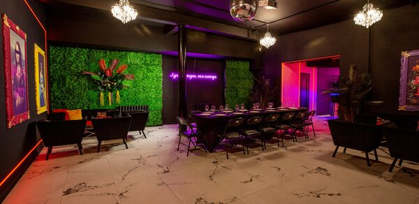 Белый неоновый лофт-зал. Торжественное пространство с зелёной растительной стеной, длинным столом и стульями. Потолок тёмный, на нём зеркальный шар и люстры. Светлые мраморные полы. На стене неоновая надпись и картины. Освещение разноцветное.
