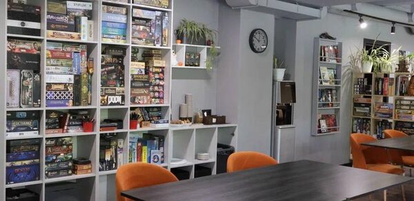 Антикафе площадью 60 м² на 30 гостей. Игровое пространство с открытой планировкой, стеллажи с настольными играми вдоль стены, серые стены и потолок, деревянные полы. Несколько столов с черными поверхностями, мягкие оранжевые стулья. Слева часы на стене, подвесное освещение, живые растения.