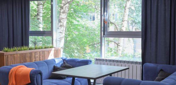 Антикафе площадью 60 м² на 30 гостей. Два синих дивана, прямоугольный стол, деревянный пуф с растениями, большие окна с видом на деревья, тёмные шторы, радиатор.