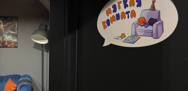 Мягкая комната на 6 гостей. Часть комнаты с серыми стенами, на стене постер. Диван синего цвета с оранжевыми подушками. Большой торшер с белым абажуром. На двери наклейка с иллюстрацией кресла и текстом «Мягкая комната».