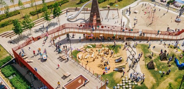 Площадка в Москве c большой фотографией. На изображении современная детская площадка с разнообразными игровыми зонами: деревянные конструкции, песочница, горки и дорожки. По площадке играют дети и гуляют взрослые, все окружено зеленью.