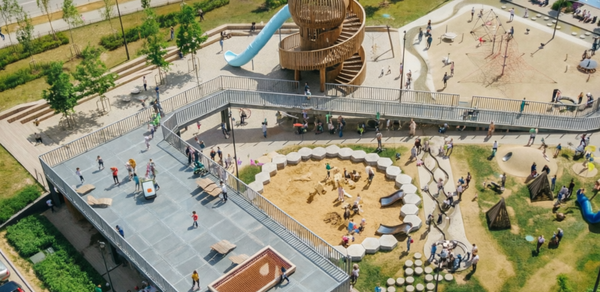 Площадка c одной фотографией в Москве. Детская площадка с деревянной башней и синим спуском, окружённая песочницей с сиденьями. Асфальтированная площадка с деревянными скамейками. Открытая местность с зелёными насаждениями и дорожками. Множество людей.