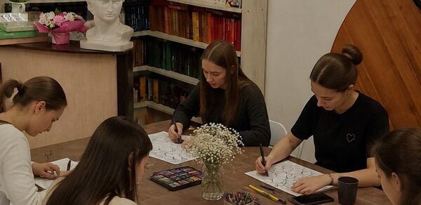 Творческая лофт-студия под мастер-классы и встречи. Комната с деревянными столами, на которых расположены цветные карандаши и блокноты. Несколько человек сидят за столом и рисуют. На заднем плане видны стеллажи с книгами, белый бюст и цветы в вазе. Светлое освещение, стены светлые.