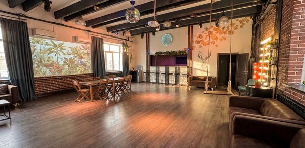 Лофт с верандой, мангалом и сценическим оборудованием. Большое помещение с деревянным полом и кирпичными стенами. Потолок с балками и подвесными светильниками. Барная стойка с высокими стульями. Диваны и обеденный стол с деревянными стульями. Декоративные элементы на стенах и зеркальный шар под потолком.