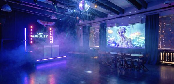 Лофт с верандой, мангалом и сценическим оборудованием. Праздничное пространство в стиле лофт с деревяными балками на потолке и паркетным полом. DJ-стойка с неоновыми огнями и надписью. Зал с деревянными столами и стульями. Дискобол и проекция на стене. Мягкое разноцветное освещение.