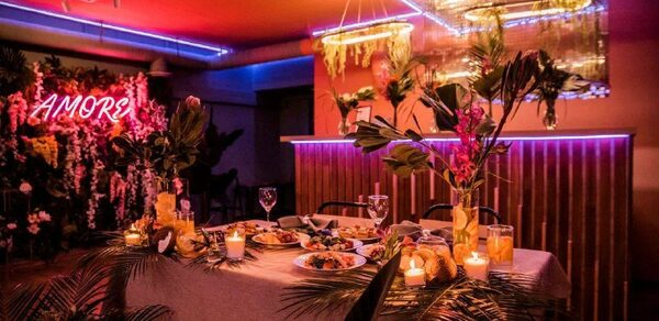 Тропический лофт с современным дизайном и верандой. Праздничное пространство с сервированным столом на шесть персон, украшено зеленью и свечами. Неоновая надпись "AMOR", яркое освещение синими и красными тонами, подвесные светильники. Стены с деревянными панелями, потолок украшен зеркальной плиткой.