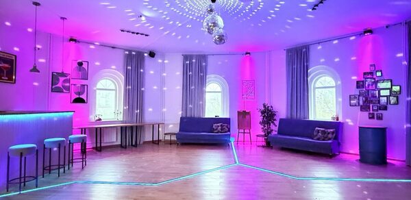 Центрум. Уютное светлое пространство круглой формы. Просторная комната с деревянным полом, мягкой мебелью в виде диванов, барной стойкой и стульями. Округлые окна с шторами. Декор дополняют растения и картины. Освещение разноцветное с акцентом на розовый, есть диско-шары. Стены белые.