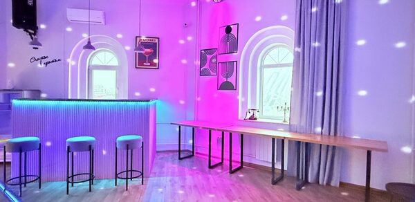 Центрум. Уютное светлое пространство круглой формы. Барная зона с подсветкой, высоким столом и тремя стульями. Деревянный пол, белые стены с арочными окнами. Подвесные светильники, дискотечное освещение. Длинный стол у стены, постеры, серые занавески, кондиционер на стене.