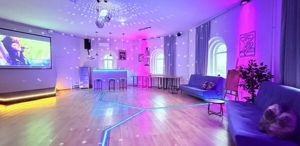 Центрум. Уютное светлое пространство круглой формы. Просторное помещение с диджейским пультом и экраном, деревянный пол, неоновая подсветка, дискошар на потолке, два синих дивана, арочные окна, высокий стол с табуретами, цветные светильники, украшения на стенах, растения.
