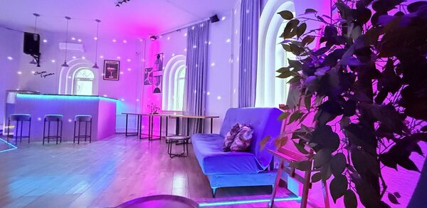 Центрум. Уютное светлое пространство круглой формы. Пространство с фиолетовой подсветкой, барная стойка с высокими стульями, деревянный пол, диван с подушками, столики, арочные окна с занавесками, декоративное растение, подвесные светильники, потолочный свет.