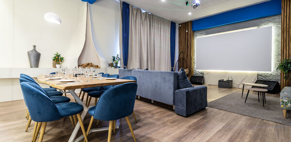 Rubin Loft (Рубин Лофт). Зал Сицилия. Современное праздничное пространство в светлых тонах с деревянным полом. Большой обеденный стол с синими стульями. Зона отдыха с серым диваном. Декоративное освещение, висящая лампа, проектор, занавески, декоративные элементы, растения.