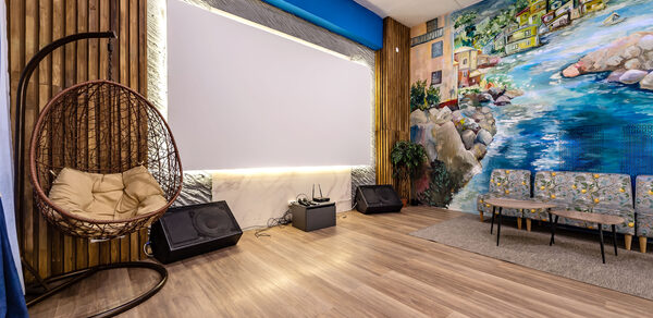 Rubin Loft (Рубин Лофт). Зал Сицилия. Помещение с деревянным полом, висячим плетеным креслом, светлой стеной с крупным экраном, двумя динамиками, мягким диваном. Одна стена украшена ярким настенным рисунком с изображением побережья.