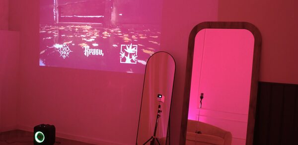 Стильный лофт для уютных вечеринок. Проекционный экран на стене с изображениями и текстом. Напольное зеркало с деревянной рамой. Колонка на полу. Розовое освещение, паркетный пол, светлый диван за углом. Камера на штативе.