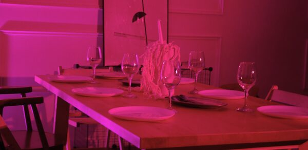 Стильный лофт для уютных вечеринок. Прямоугольный деревянный стол с расставленными белыми тарелками и бокалами для вина. В центре стола декоративная композиция. Окружают разностильные стулья. Освещение розового цвета, стены с белыми панелями.