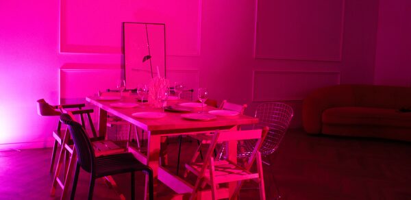 Стильный лофт для уютных вечеринок. Современное пространство с длинным деревянным столом, окруженным различными стульями, на столе посуда. Яркое розовое освещение. На стене панели, в углу диван. На столе зеркало. Пол паркетный.