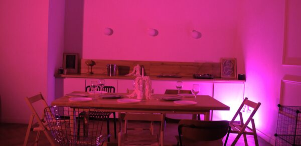 Стильный лофт для уютных вечеринок. Столовая зона с деревянным столом, окружённым разнообразными стульями. На столе белая салфетка и бокалы. Позади небольшая барная стойка с предметами декора и освещением розового тона. Пол паркетный, на стене три светильника.