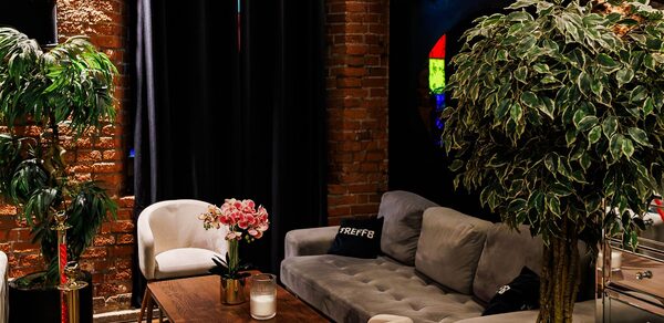 Двухуровневая event-площадка 550 м². Лофт-пространство с кирпичными стенами и темными шторами. Мягкий серый диван и кресла вокруг деревянного стола. Декоративная растительность, свеча на столе, мягкое освещение.