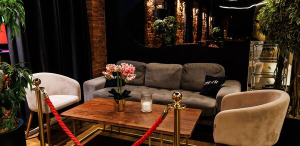 Двухуровневая event-площадка 550 м². Лаунж-зона с серым диваном и двумя белыми креслами вокруг деревянного стола, украшенного цветочным горшком и свечой. Красная веревка на золотых стойках. Зелёные растения по краям. Темные занавески и кирпичная стена. Зеркало на стене.