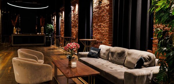 Двухуровневая event-площадка 550 м². Лофт-пространство с кирпичными стенами и деревянным полом. Мягкий серый диван, два бежевых кресла и деревянный стол с цветами и свечой. Черные занавески и круглые светильники над зоной отдыха.