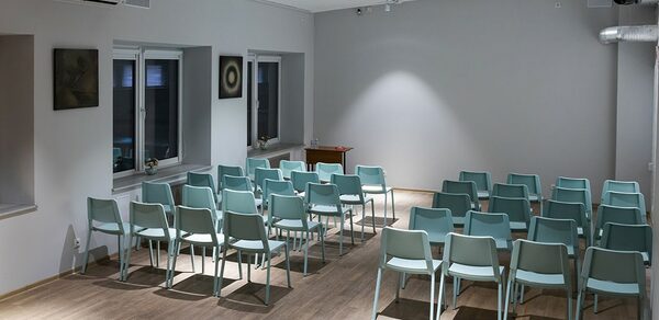 Пространство для ваших праздников. Конференц-зал с деревянным полом, расставлены ряды мятных стульев. Светлые стены, на одной из них картины. Потолочные светильники обеспечивают освещение. Есть кондиционер и окна с откосами. У стены небольшой деревянный стол.