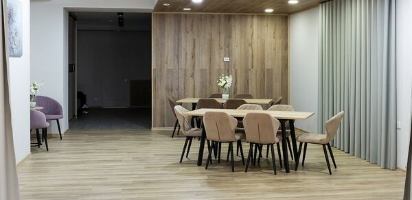 Пространство для ваших праздников. Просторное помещение с деревянным полом и панелями на стенах и потолке. Две группы столов и стульев, цветы на столах. Освещение представлено встроенными светильниками. Справа - шторы, слева - картина и пара кресел.