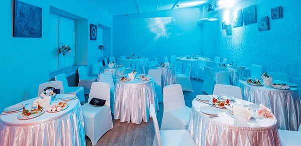 Пространство для ваших праздников. Банкетный зал с круглыми столами, покрытыми белыми скатертями, и белыми стульями. Голубое освещение. На стенах картины. Декор из цветов. Блюда с закусками на столах. Пол деревянный. Потолочное освещение.