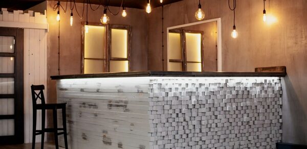 Лофт-пространство в Ярославле. Барная стойка с отделкой из белых деревянных блоков, стул, подвесные лампы с теплыми лампочками, деревянные стены с двумя окнами, пол светлого дерева, черный потолок с проводами, светодиодная подсветка стойки.