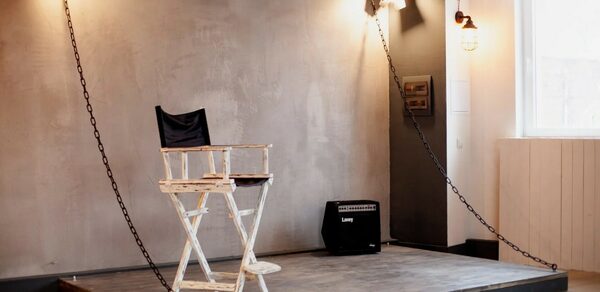 Лофт-пространство в Ярославле. Пространство с серыми стенами, черным потолком и деревянным полом. Небольшая сцена с деревянным режиссерским стулом, по бокам цепи. Освещение от двух настенных светильников и окна. Мощник стоит на сцене.