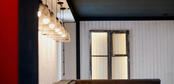 Лофт-пространство в Ярославле. Мягкий уголок коричневого цвета с вертикальным освещением на стене, деревянные панели белого цвета, в углу окно с рамой тёмного цвета. Потолок тёмный, освещение от лампочек с прозрачными плафонами вдоль стены.