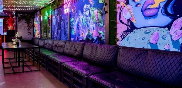 Яркий лофт для праздников. Праздничное пространство с длинным черным диваном и черными столами, яркие граффити на стенах, разноцветная светодиодная подсветка на потолке, ковёр с орнаментом на полу, стены украшены зелёными растениями, надпись "Social House" за диваном.