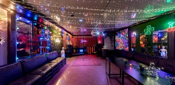 Яркий лофт для праздников. Караоке-зал с диванами вдоль стен, деревянные столы, зеркальные панели, яркие неоновые и светодиодные украшения на стенах, цветной ковёр по центру, отражения огней на потолке, тёмная цветовая гамма интерьера.