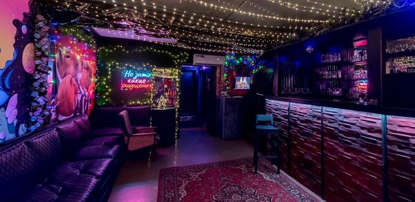 Яркий лофт для праздников. Клубное пространство с черным кожаным диваном, барной стойкой, полками с напитками. Потолок украшен гирляндами, стены с неоновыми знаками и граффити. На полу ковёр с орнаментом. Освещение приглушенное, неоновое и гирляндное.