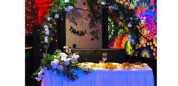 Яркий лофт для праздников. Светлое помещение с декорированным столом, покрытым белой скатертью, украшенным цветочной аркой. На столе еда и напитки. На стене яркие рисунки и надпись "Танцы!" Окружение подсвечено разноцветными огнями.