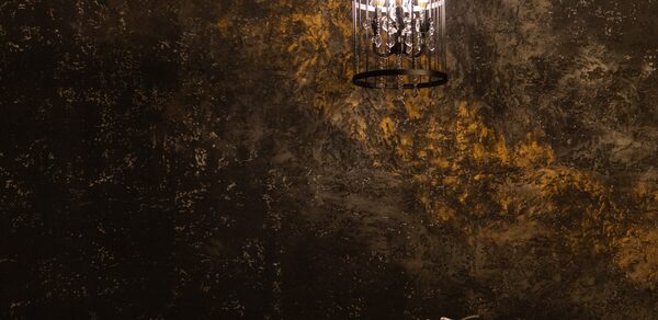 Лофт со своим двориком. Потолочный светильник в виде клетки подвешен на фоне текстурированной темной стены с золотистыми акцентами. Справа внизу металлический декоративный элемент с абстрактным узором. Освещение теплое.