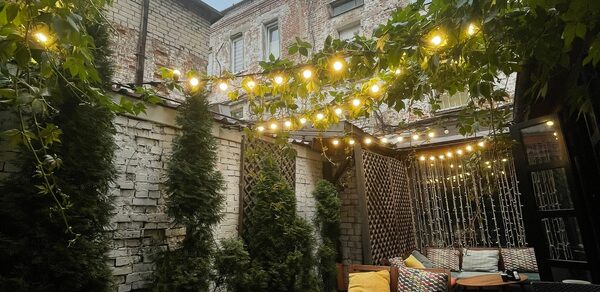 Лофт со своим двориком. Внутренний двор с деревянными столами и плетеными стульями с подушками. На стенах кирпич и зелень. Над пространством висят гирлянды с лампочками. В углу живая изгородь. На полу плитка. Стены окружающих зданий из кирпича.