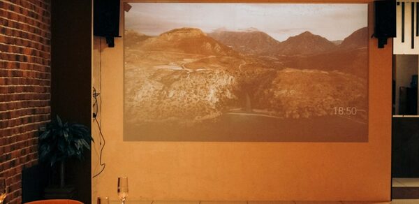 Антикафе с несколькими залами. Помещение с кирпичной стеной, проектором на гладкой стене, демонстрирующим горный пейзаж. Слева стол с бокалами и тарелками. Оранжевое освещение, белые плиточные полы, рядом с экраном небольшая растительность. Справа вход в другую зону.