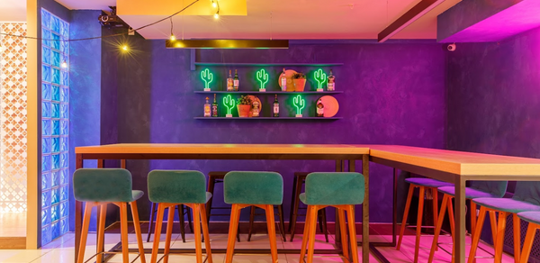 Антикафе с несколькими залами. Барная зона с высокими деревянными столами и барными стульями с бирюзовой обивкой. Темно-синие стены с неоновыми вывесками в виде кактусов. Подвесное освещение и настенная полка с декоративными элементами. Плиточный пол.