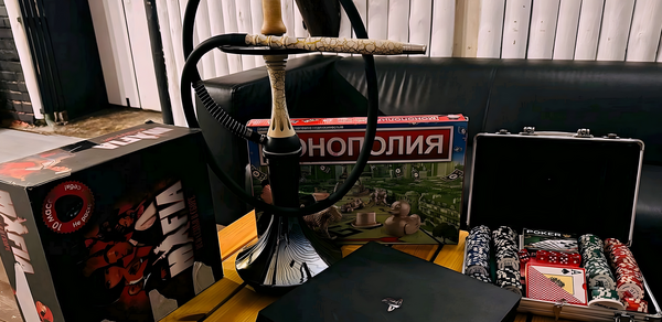 Лофт-мансарда . Кофейный столик из светлого дерева с кальяном, настольные игры «Мафия» и «Монополия», набор для покера с фишками и картами в открытом кейсе. На заднем плане черный кожаный диван и белая деревянная обшивка стен.