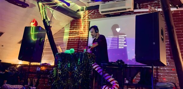 Лофт-мансарда . Лофт-пространство с кирпичными стенами и деревянными балками. Диджей за пультом, украшенным гирляндами и тканью. Справа и слева установлены крупные колонки. На стене проектор с текстом и изображением сердца. На потолке кондиционер. Цветное освещение.
