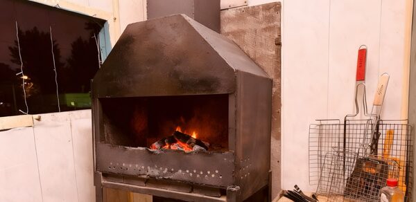 Лофт-мансарда . Камин в металлическом корпусе с дровами, белые деревянные стены, тёмное квадратное окно слева, рядом с камином инструменты, сетка для гриля, деревянная поверхность. Освещение тёплое.