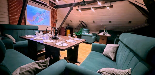 Лофт-мансарда . Лофт с деревянными балками, кирпичными стенами и наклонным потолком. Угловой диван и кресла бирюзового цвета, деревянный стол с сервировкой для ужина. Проектор с экраном, подвесные светильники, подвесное кресло. Светлое деревянное покрытие пола.