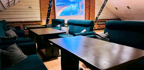 Лофт-мансарда . Лофт с кирпичной стеной и белым наклонным потолком. Мягкие тёмно-зелёные диваны и деревянные столы. Настенный проектор показывает изображение. Декоративные светодиодные гирлянды. Светлое деревянное покрытие пола.