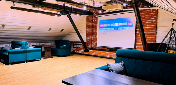 Лофт-мансарда . Лофт-пространство с деревянным полом и кирпичными стенами. Мягкие диваны зелёного цвета, деревянные балки на потолке с точечным освещением. Проекционный экран на стене. Цвет стены белый с серыми элементами.