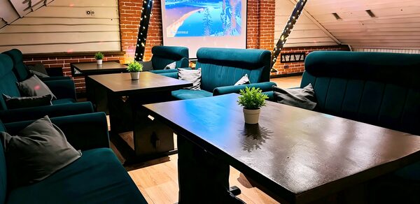 Лофт-мансарда . Лофт с кирпичными стенами и деревянными панелями. Цифровой экран на стене, темные деревянные столы, темно-зеленые кресла с подушками. На столах горшки с зелеными растениями. Зафиксировано декоративное освещение. Светлый деревянный пол.