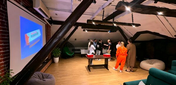 Krisha loft (Крыша Лофт). Лофтовой пространство с деревянным полом и кирпичными стенами. На стене висит большой экран. Мебель: диваны, кресло-мешок, стол с плоской столешницей. Несколько человек стоят у стола. Освещение подвесными лампами, интерьер выдержан в спокойных тонах.