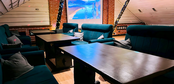 Лофт-мансарда . Чердачное помещение с кирпичными стенами, деревянные столы и мягкие кресла темно-зеленого цвета, подушки серого оттенка. На стене проектор с изображением зимнего пейзажа. Потолок с деревянными балками и гирляндами. Пол деревянный.