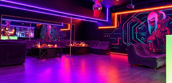Cyberpunk (Киберпанк). Клубное помещение с темными стенами и неоновой подсветкой, мягкая мебель по периметру, зеркала на стенах, световые панели с геометрическим рисунком, деревянный пол. Центральная часть украшена столиками с напитками и свечами.
