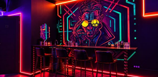 Cyberpunk (Киберпанк). Барная стойка с высокими стульями, стена с неоновым граффити, подсветка в фиолетовых и розовых тонах, бокалы и бутылки на стойке, тёмные стены, зеркало, пол из тёмного дерева, яркое освещение.