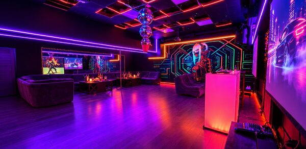 Cyberpunk (Киберпанк). Клубное помещение с неоновым освещением, фиолетовыми и красными акцентами. Зона отдыха с темными диванами и столами, на стенах графические изображения. Потолок с квадратными панелями, светодиодные лампы, дискошар подвешен к потолку.